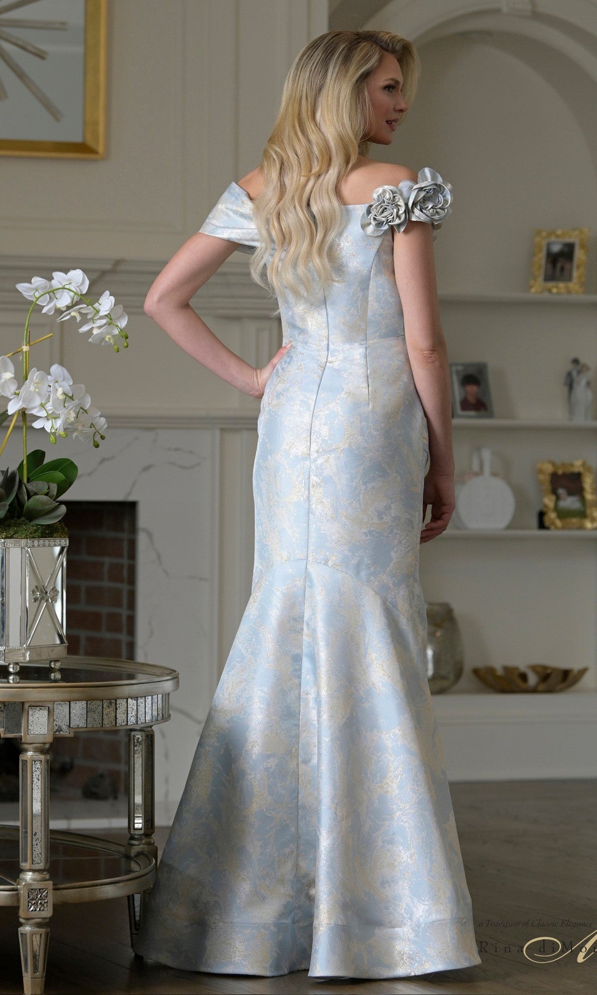 A woman with long blonde hair wears the Rina Di Montella RD3522, a light blue and silver jacquard off-the-shoulder gown with floral accents, standing in a softly lit, elegant living room.
